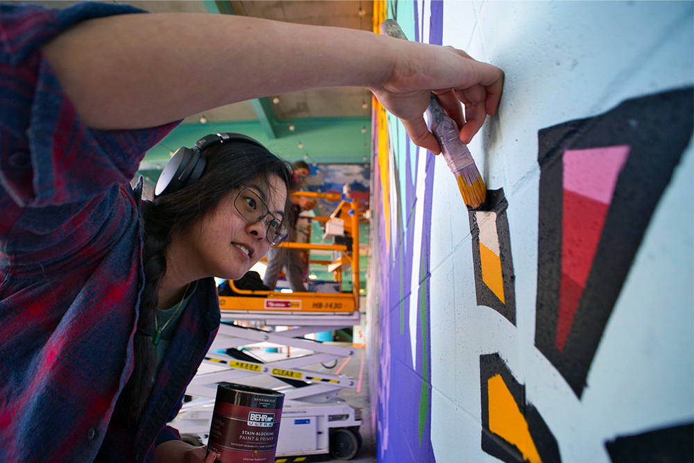 A young woman wearing headphones and glasses leaning in to look at the mural she is painting