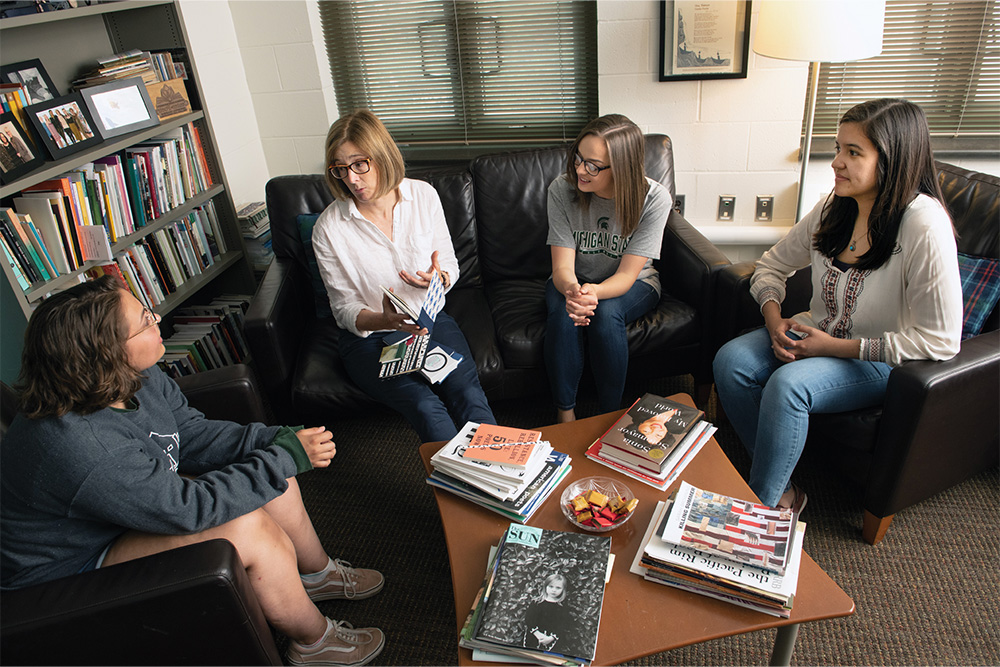 Four women having a discussion while sitting in dark leather chairs next to a coffee table full of books