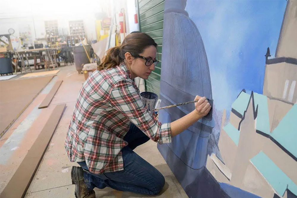 Woman with glasses and plaid shirt kneeling while painting scenery for a theatre perforance